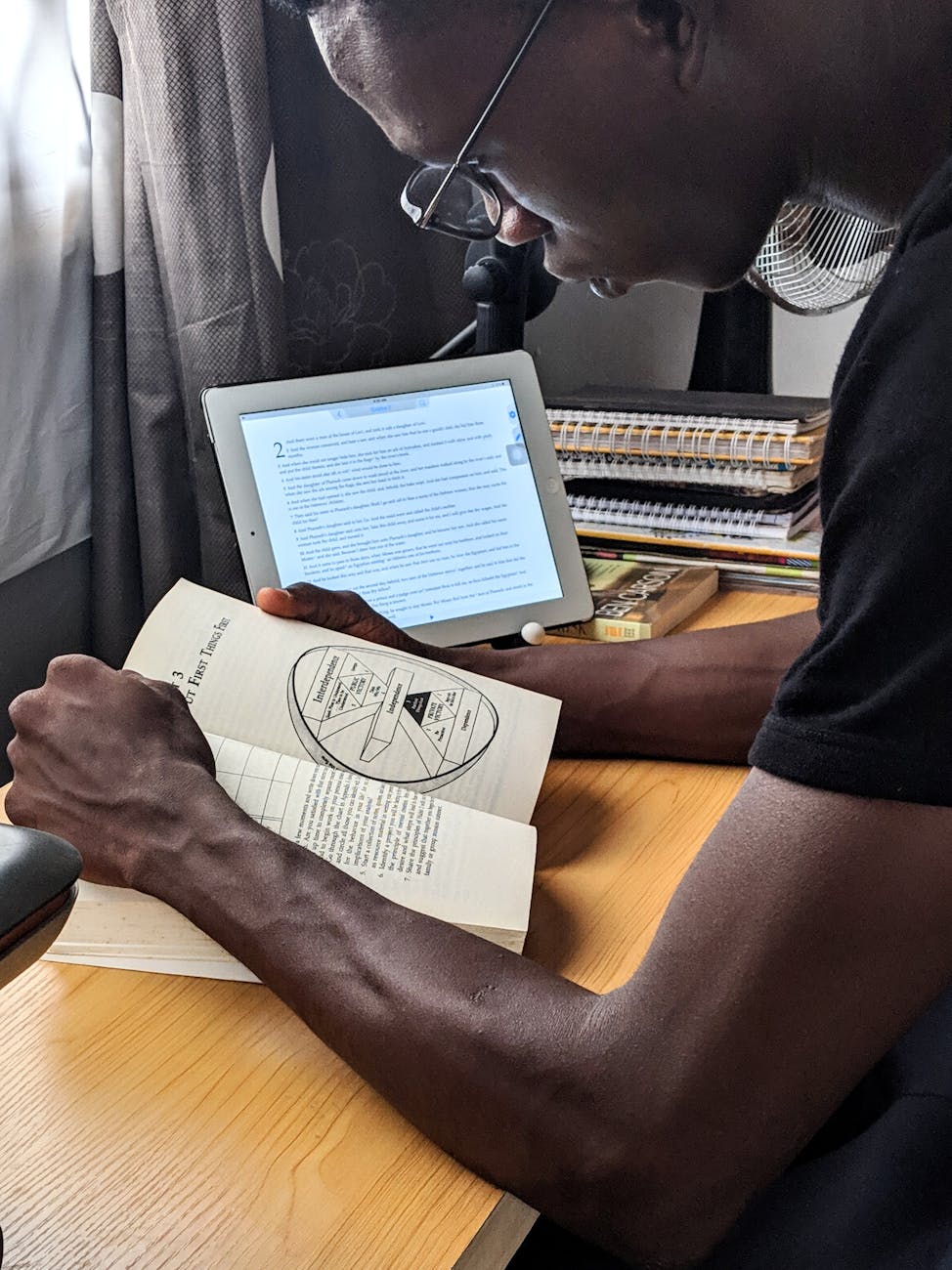 Study Abroad A man studying with a book and tablet at a desk indoors, showcasing multitasking and modern study habits.