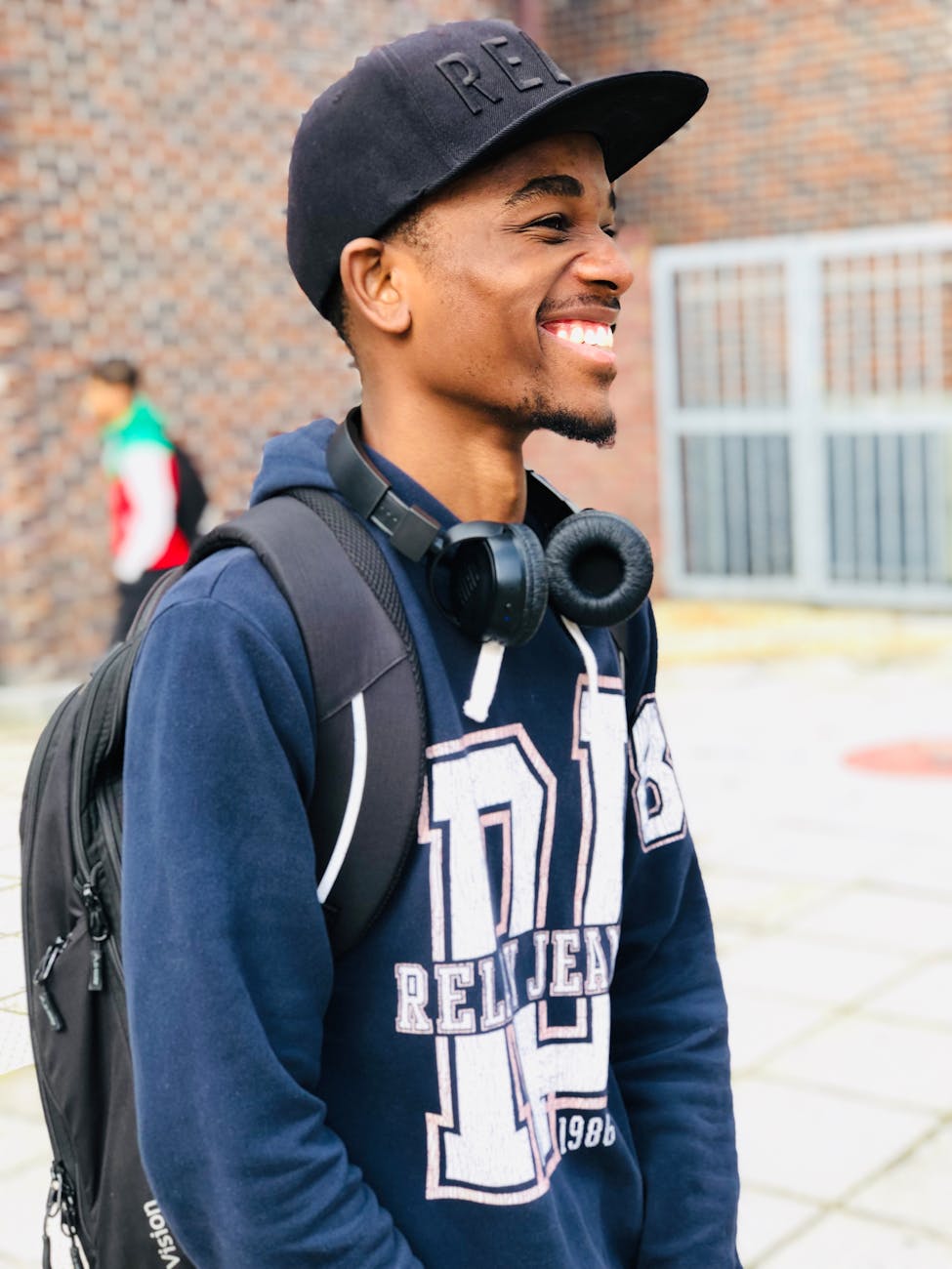 Study Abroad Smiling young man wearing cap and headphones, standing with backpack outdoors.